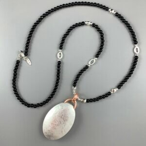 Long black bead necklace interspersed with silver beads and silver oval links, with a large solid oval pendant hanging from a rose gold hook